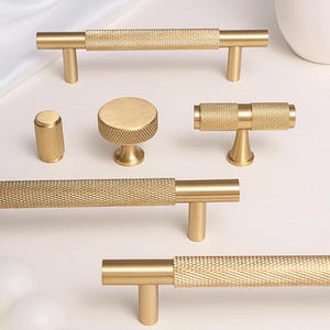 Op de afbeelding: Een verzameling goudkleurige kastbeslagen, waaronder stanggrepen, knoppen en een cilindrische greep. De hardware heeft een textuur, diamantvormig ontwerp. De stukken zijn gerangschikt op een witte ondergrond.