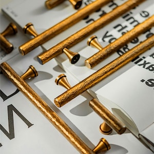 Puede incluir: Primer plano de varios tiradores de armario dorados con textura martillada. Los tiradores están dispuestos sobre una superficie blanca con texto. La imagen también incluye una pluma y el texto "Prprfctn Treasures".