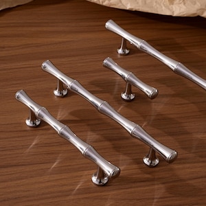 May include: Silver-toned cabinet pulls with a bamboo-inspired design. The pulls are arranged on a wooden surface, showcasing their polished finish and decorative appeal. The image includes the text "Prprfctn Treasures".