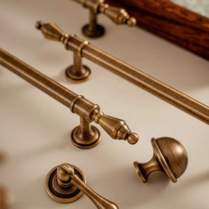 May include: Close-up of several antique bronze cabinet hardware pieces. The collection includes long bar pulls, a knob, and a handle with a round backplate. The hardware has a detailed, textured finish and a warm, aged appearance.