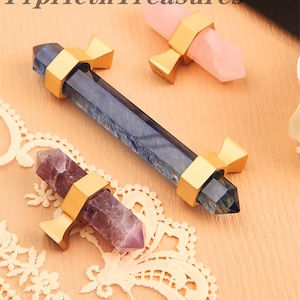 Op de afbeelding: Drie edelsteen kristallen toverstokken met goudkleurige metalen accenten. De kristallen zijn in tinten blauw, roze en paars. De toverstokken worden getoond op een lichtgekleurd oppervlak met een witte kanten rand.