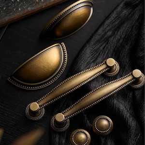 May include: A selection of antique brass cabinet hardware, featuring two cup pulls, two bar pulls, and two round knobs. The hardware has a detailed design with a warm, aged finish, ideal for adding a vintage touch to furniture. The text "PrprfctnTreasures" is visible at the top.