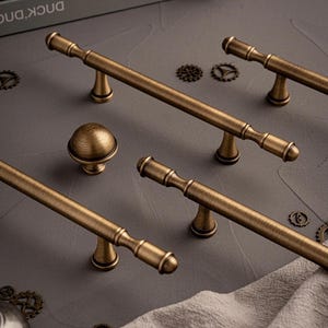 Könnte beinhalten: Eine Auswahl an antiken Messing-Schrankbeschlägen, darunter mehrere Stangengriffe und ein runder Knauf. Die Beschläge haben eine Vintage-Ästhetik mit einem warmen, gealterten Finish. Die Teile sind auf einer grauen Oberfläche angeordnet.