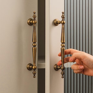 Peut inclure: Poignées de placard en laiton antique avec des détails ornés. Les poignées sont montées verticalement sur une porte d'armoire blanche. Le texte "Prprfctn Treasures" est en haut de l'image.