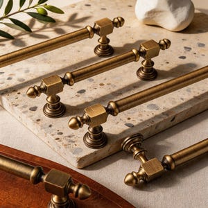May include: Close-up of several antique brass cabinet pulls. The pulls have a rectangular center and rounded ends. The hardware is displayed on a speckled stone surface, with a wooden tray and olive branch in the background.