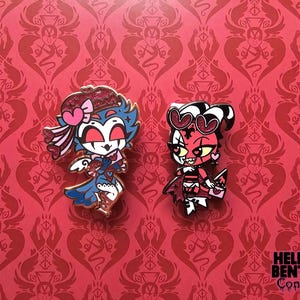 以下が含まれることがあります： 漫画のキャラクターが描かれた2つのエナメルピン。左のピンは、ピンクのハートとグリッターのアクセントが付いた青と白のフクロウのようなキャラクターです。右のピンは、ハート型の目とグリッターのディテールが付いた赤と黒の小鬼のようなキャラクターです。