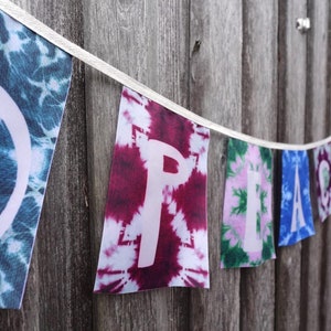 Könnte beinhalten: Ein Banderole aus Batikstoff mit dem Wort "FRIEDEN" in weißen Buchstaben. Jeder Buchstabe befindet sich auf einer separaten Flagge mit einem anderen Batik-Muster. Die erste Flagge hat ein Friedenszeichen darauf.