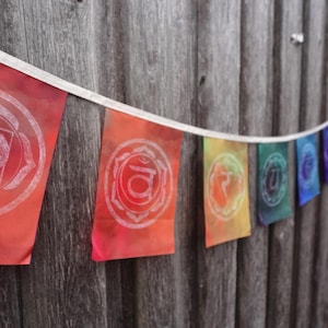 May include: A string of seven colorful flags with white symbols representing the seven chakras. The flags are hanging on a wooden fence.