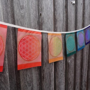 Peut inclure: Une guirlande de drapeaux en tissu colorés avec un motif géométrique au centre de chaque drapeau. Les drapeaux sont dans des tons de rouge, orange, jaune, vert, bleu et violet.