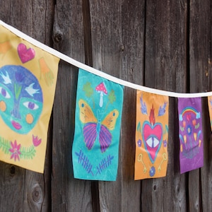 Könnte beinhalten: Ein farbenfrohes Stoffbanner mit einer Schnur von Flaggen. Jede Flagge zeigt ein anderes Design, darunter ein Gesicht, ein Schmetterling, ein Herz und ein Regenbogen. Die Flaggen sind an einer weißen Schnur befestigt und hängen vor einem hölzernen Hintergrund.