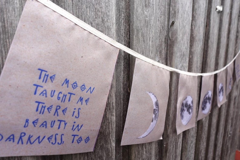 K&ouml;nnte beinhalten: Ein Banner mit braunen Papierfahnen, die mit wei&szlig;em Garn zusammengef&uuml;gt sind. Die Fahnen zeigen schwarz-wei&szlig;e Bilder des Mondes in verschiedenen Phasen. Eine Fahne tr&auml;gt einen blauen Text, der lautet: "Der Mond hat mir beigebracht, dass es auch in der Dunkelheit Sch&ouml;nheit gibt."