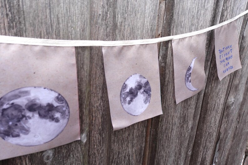 K&ouml;nnte beinhalten: Eine Schnur aus Stofffahnen mit verschiedenen Mondphasen und dem Text "That moon doesn't belong to me, but I am loved".