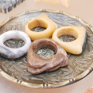 Puede incluir: Una colección de anillos en varios colores y diseños, incluyendo un anillo blanco veteado, un anillo marrón y dos anillos amarillos. Los anillos se exhiben en un plato decorativo, redondo, de color bronce con un patrón de hojas.
