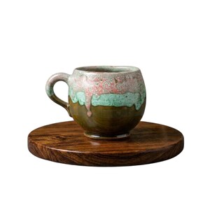 Puede incluir: Una taza de cerámica hecha a mano con un esmalte único en tonos verdes, rosas y marrones, colocada sobre un posavasos redondo de madera. La taza tiene una forma redondeada con un asa. El posavasos es de madera oscura.