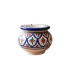 Puede incluir: Un pequeño cenicero redondo de cerámica con borde azul e interior blanco. El exterior presenta un patrón repetitivo de diseños florales azules, rojos, verdes y naranjas sobre un fondo crema. El cenicero tiene una pequeña abertura circular.