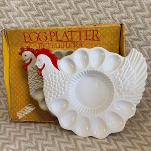 Puede incluir: Un plato para huevos de cerámica blanca con forma de gallina, con cresta y barbillas rojas, que presenta hendiduras circulares para huevos. El plato se presenta en una caja de cartón amarilla con el texto EGG PLATTER BY TELEFLORA en rojo.