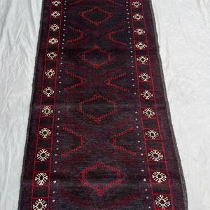 Pode incluir: Um tapete retangular de cor escura com um padrão geométrico. O tapete apresenta um design central de formas de diamante em vermelho, rodeado por uma borda de motivos florais vermelhos e brancos. O tapete tem uma borda com franjas.