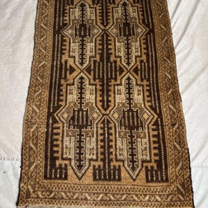 May include: A rectangular woven rug featuring a geometric pattern in shades of dark brown, tan, and beige. The design includes symmetrical, stepped, and angular motifs throughout the center, framed by a decorative border and short fringe at both ends.