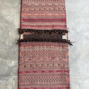 May include: A rectangular, hand-woven rug with a complex pattern of red, brown, and tan stripes and geometric shapes. The rug has a fringed edge and a central section with a darker brown, textured design. The rug is displayed on a gray surface.