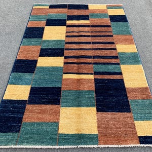 Op de afbeelding: Een rechthoekig vloerkleed met een geometrisch patroon van afwisselende blokken en horizontale strepen in marineblauw, mosterdgeel, petrolgroen en terracottabruin, afgewerkt met een korte witte franje aan de onderkant.
