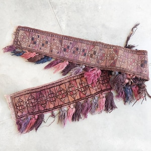 May include: Two decorative textile bands with geometric patterns in shades of burgundy, pink, and brown. Each band is adorned with colorful tassels in varying shades of pink, purple, and blue. The bands appear to be handmade.