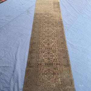 May include: A long, rectangular rug with a detailed, geometric pattern in shades of brown and beige. The rug has a fringe on one end and is likely made of woven material. The design features repeating floral and geometric motifs, creating a traditional aesthetic.