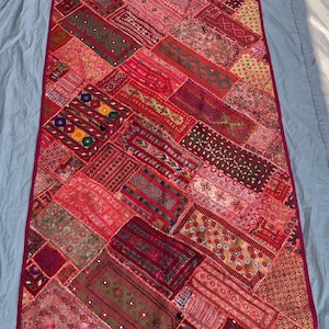 May include: A rectangular patchwork textile in shades of red, pink, and burgundy. The design features various geometric and floral patterns, with a deep red border. The textile is likely a decorative item, such as a wall hanging or throw.