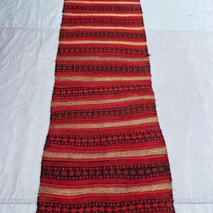 May include: A long, rectangular runner rug featuring a repeating pattern of horizontal stripes in shades of deep red, black, and tan. The design includes intricate geometric motifs woven into the darker bands, with fringed edges at both ends.