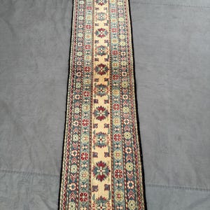 Puede incluir: Una alfombra de pasillo rectangular con un campo central beige y intrincados diseños florales en rojo, azul y verde. La alfombra tiene un borde negro y bordes con flecos, mostrando un diseño tradicional. La alfombra mide aproximadamente 60 cm de ancho.