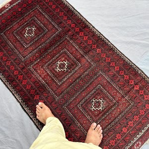 Può includere: Un tappeto rettangolare, tessuto a mano, con una combinazione di colori rosso scuro e marrone. Il tappeto presenta un motivo geometrico ripetitivo di quadrati e diamanti, con dettagli intricati in bianco e nero. I bordi del tappeto sono rifiniti con frange.