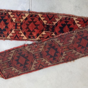 Op de afbeelding: Twee antieke, smalle, geweven tapijten met geometrische patronen. De tapijten zijn voornamelijk rood met zwarte en bruine accenten. Het ontwerp bevat herhaalde ruit- en driehoekvormen. De tapijten worden gepresenteerd op een lichtgrijze ondergrond.