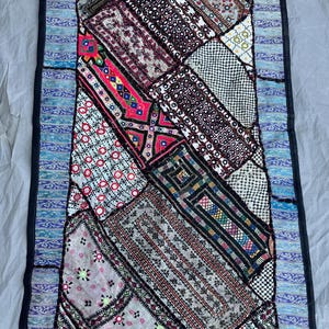 May include: A rectangular patchwork textile with a variety of patterns and colors. The central section features a mix of geometric and floral designs in shades of red, pink, black, and white. A blue border with a decorative pattern surrounds the central panel.