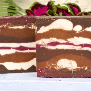 May include: Two bars of handmade soap with a layered design. The soap is brown, white, and red. The soap is on a white surface.