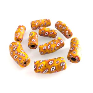May include: A collection of cylindrical beads with a yellow base color and a floral pattern in red, white, and black. The beads are scattered on a white surface. These beads are suitable for jewelry making.