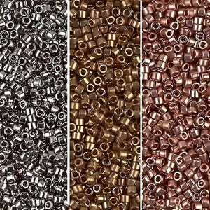 Puede incluir: Primer plano de tres secciones de pequeñas cuentas metálicas. La sección izquierda presenta cuentas plateadas, la sección central tiene cuentas doradas y la sección derecha muestra cuentas de color cobre. Estas cuentas son cilíndricas con un agujero en el centro.