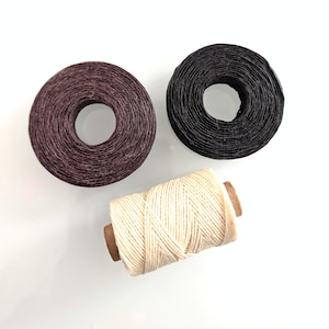 May include: Three spools of thread are arranged together. Two spools are circular and flat, one in a deep dark purple and one in black. The third spool is cylindrical, positioned horizontally, and features a light cream-colored thread.