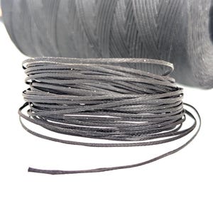 May include: Close-up of a spool of black waxed cord, tightly wound in a coil. The cord is a dark, matte black, with a slightly textured surface. A larger spool of the same cord is visible in the background, suggesting a craft or jewelry-making supply.