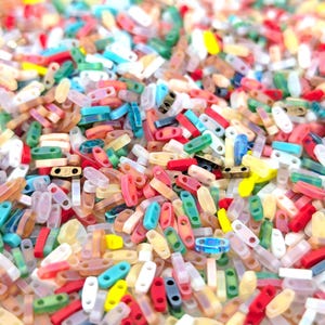 Könnte beinhalten: Nahaufnahme eines Haufens bunter, kleiner, rechteckiger Perlen. Die Perlen sind in verschiedenen Farben gehalten, darunter Rot, Blau, Gelb, Grün und Transparent. Jede Perle hat zwei Löcher. Die Perlen sind verstreut und bilden eine strukturierte Oberfläche.