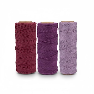 May include: Three spools of colored cotton twine in shades of burgundy, purple, and lavender. Each spool is tightly wound and mounted on a cardboard core. The twine is suitable for various crafting projects.