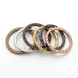 May include: A set of six circular rings featuring a hammered, textured surface. The rings are crafted in various metallic finishes, including antique brass, silver, copper, gold, dark gunmetal, and rose gold, arranged in an overlapping row.