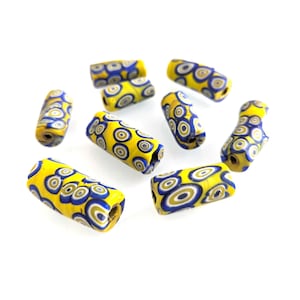 May include: A collection of yellow, blue, and white glass beads with a cylindrical shape. Each bead features a pattern of concentric circles in blue and white against a yellow background. The beads are scattered on a white surface.