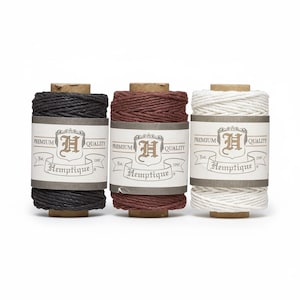 May include: Three spools of hemp twine in black, burgundy, and white. Each spool has a cardboard core and a label with the text "Premium Quality Hemptique Est. 1997". The twine is tightly wound and ready for crafting.