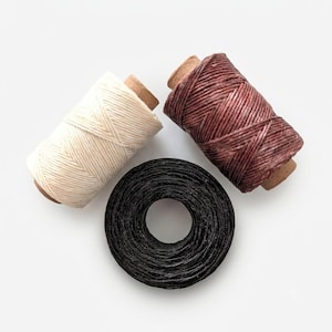 Peut inclure: Trois bobines de fil ciré disposées en formation triangulaire. Une bobine est de couleur crème, une autre est d'un brun rougeâtre profond, et la troisième est une bobine plate et circulaire de fil noir. Chaque bobine présente une texture serrée.