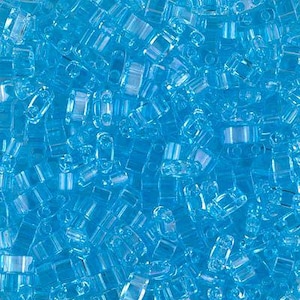 May include: A collection of small, transparent, light blue glass beads. Each bead features a rectangular shape with two distinct holes running through the center, creating a uniform, translucent appearance across the entire group of craft supplies.