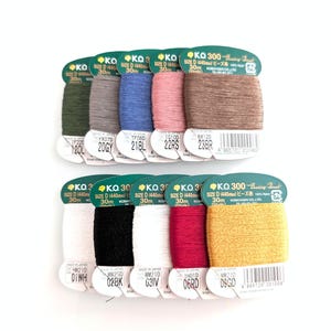 Pode incluir: Uma coleção de carretéis de linha em várias cores, incluindo verde, cinza, azul, rosa e marrom, dispostos em duas fileiras. Cada carretel é rotulado com um código de cor e o texto "KO 300". A linha tem 30m de comprimento.