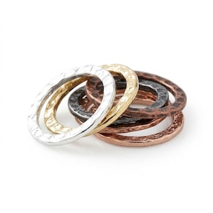 May include: A set of four stacked rings featuring a hammered, textured surface. The collection includes one silver-toned ring, one gold-toned ring, one dark charcoal-toned ring, and one copper-toned ring, all arranged in a circular, overlapping cluster.