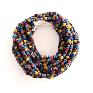 Puede incluir: Un collar de múltiples hilos compuesto por numerosas cuentas pequeñas y redondeadas en tonos vibrantes de azul, amarillo, rojo, verde y marrón, con algunas cuentas que presentan patrones de rayas blancas y negras, unido por un cordón de ganchillo blanco.