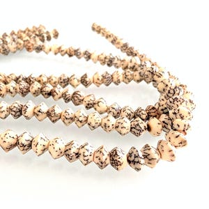 May include: Close-up of several strands of beads. The beads are a light beige color with dark brown speckled patterns. The beads are diamond-shaped and strung together, creating a necklace or bracelet.