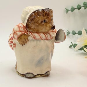 May include: A ceramic hedgehog figurine wearing a white bonnet and a white apron over a red and white checkered shirt. The hedgehog has brown fur, a black nose, and is holding a small grey object in one hand while carrying a red floral bag.