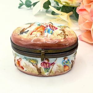 May include: Oval ceramic trinket box with a hinged lid. The lid features a pastoral scene with figures and animals. The sides of the box depict figures in a landscape. The box has a brown rim and a brass clasp. The box is decorated with pastel colours.